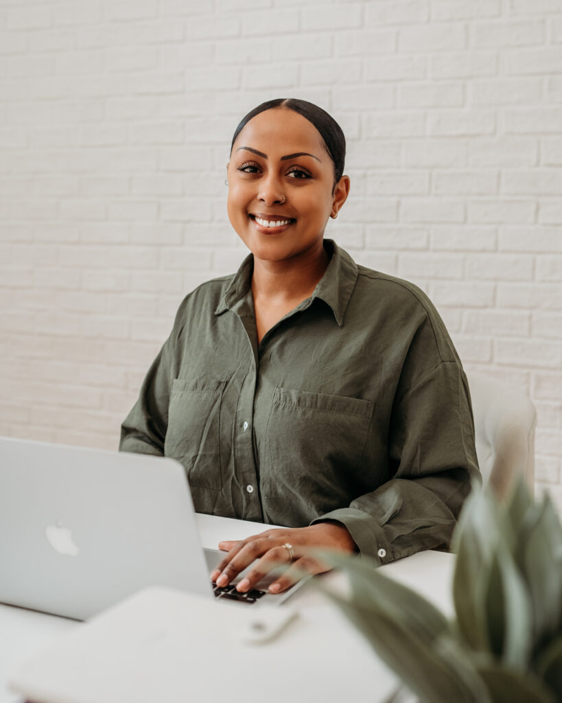 A woman sits at a desk with a laptop, smiling warmly at the camera. She is wearing a green button-up shirt and is positioned against a white brick wall, creating a clean and professional atmosphere.