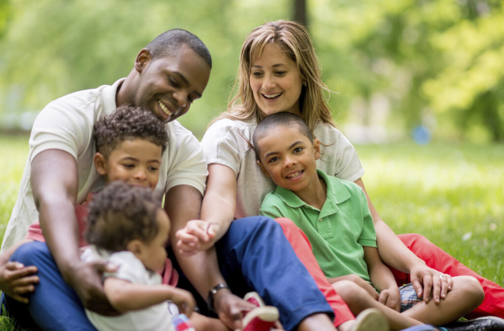 A smiling family of five sits together on the grass in a park, enjoying a sunny day. The parents and three children share a joyful moment, with greenery and trees in the background.